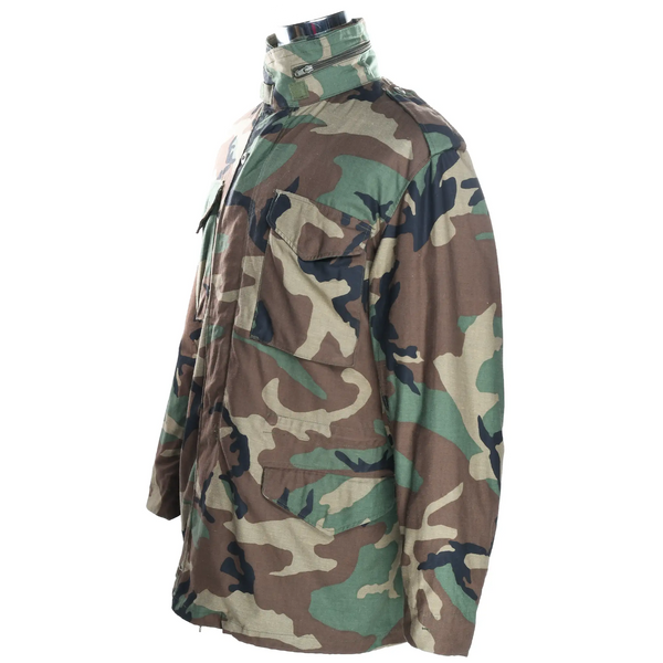 Angled front and side view of US Army M65 field jacket in woodland camo with flap pockets and stand collar.